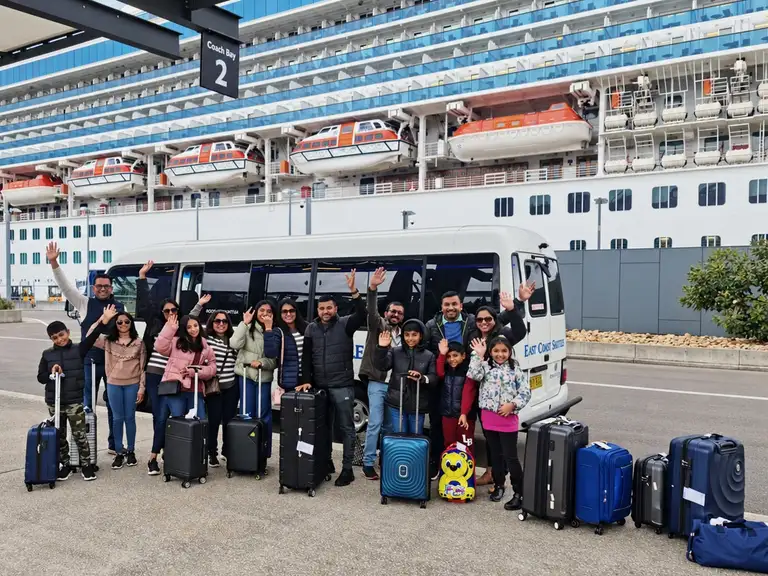 A family with their luggage waves beside a white minibus at a cruise terminal.