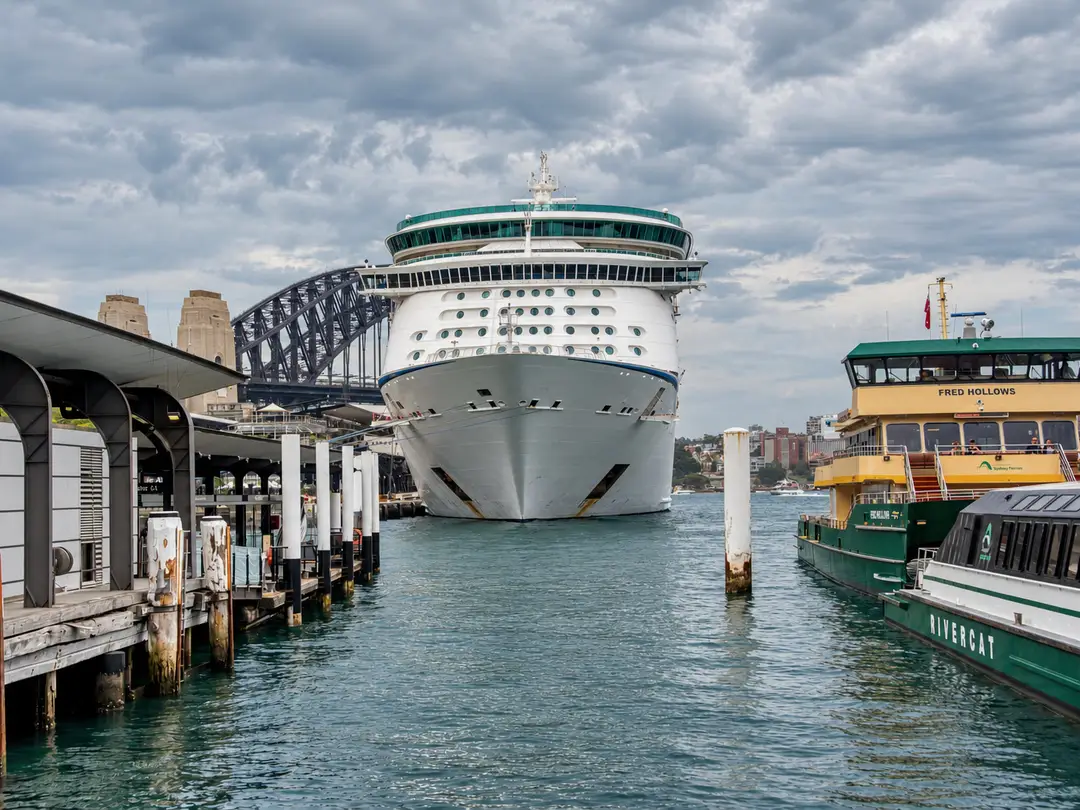 The bow of a cruise ship at a Sydney Harbour wharf, with the Harbour Bridge behind and ferries alongside under grey cloud.