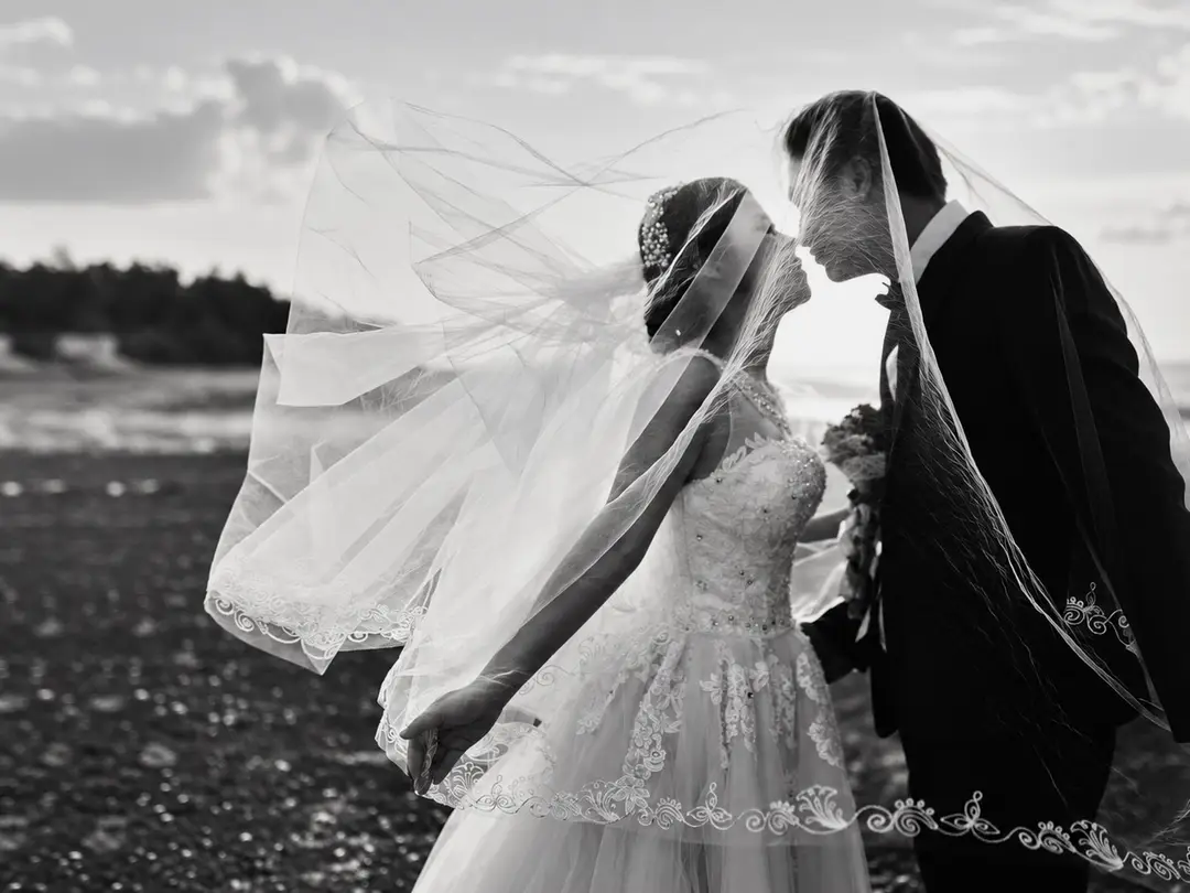 A black and white photo of a bride and groom kissing under the bride's veil outdoors, near the water.