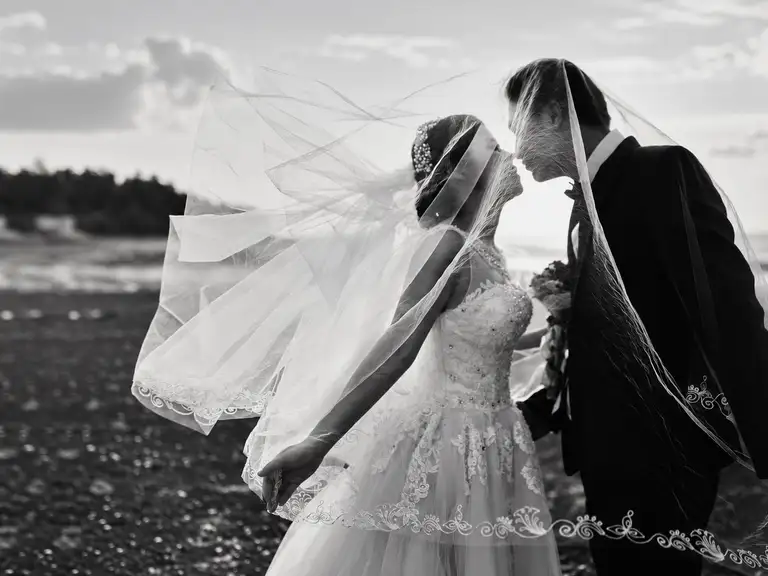A black and white photo of a bride and groom kissing under the bride's veil outdoors, near the water.